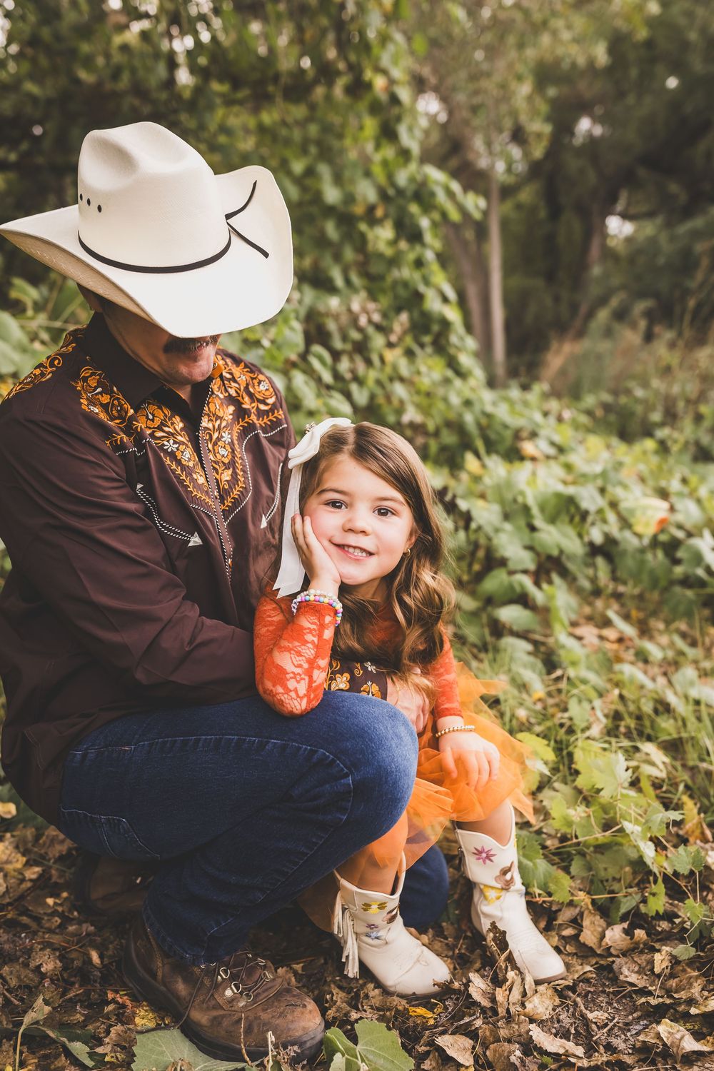 A man in a cowboy hat crouches beside a smiling young girl in cowboy boots and orange dress outdoors.