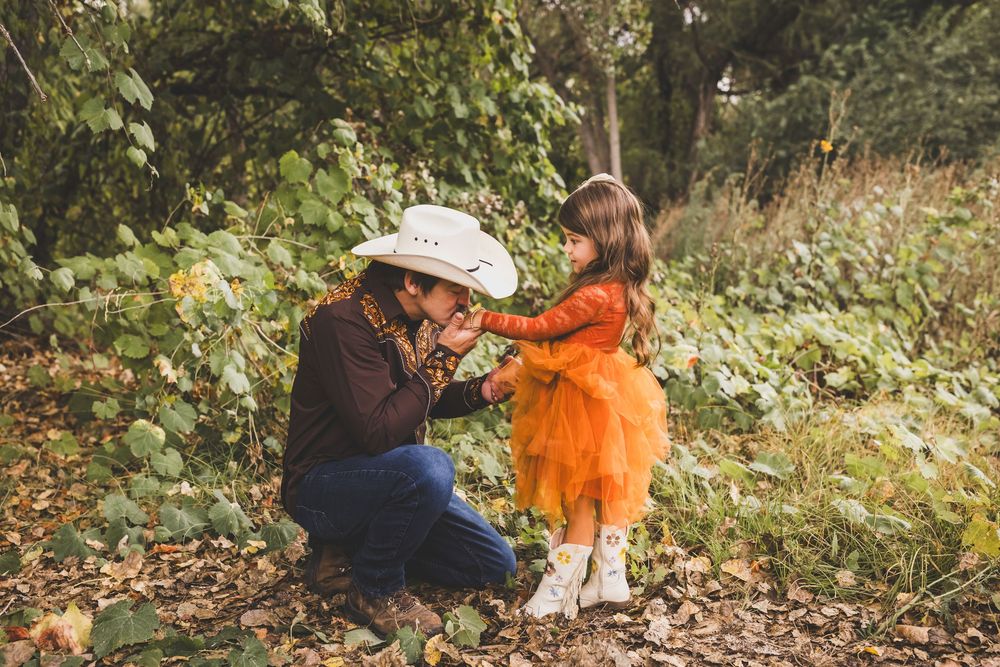 A woman in a cowboy hat crouches to interact with a young girl in an orange tutu dress outdoors.