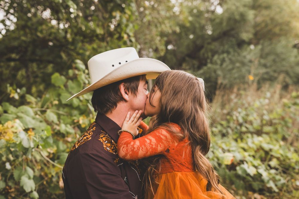 A couple shares a romantic kiss outdoors, the man wearing a white cowboy hat and brown jacket, woman in orange dress.