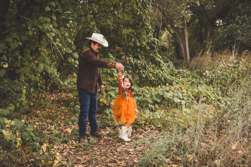 Adult in cowboy hat holds child in orange Halloween costume on a leaf-covered forest path in autumn.