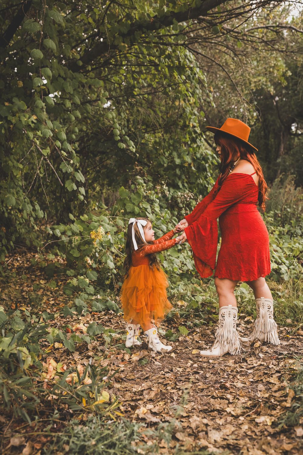 A woman in a red dress and hat holds hands with a young girl in an orange tutu in an autumn forest setting.
