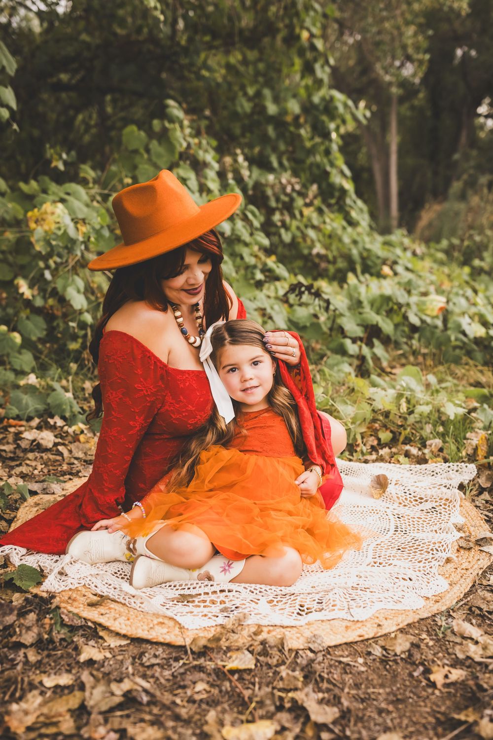 A mother in a red dress and orange hat sits with her young daughter in an orange tulle skirt on a lace blanket outdoors.