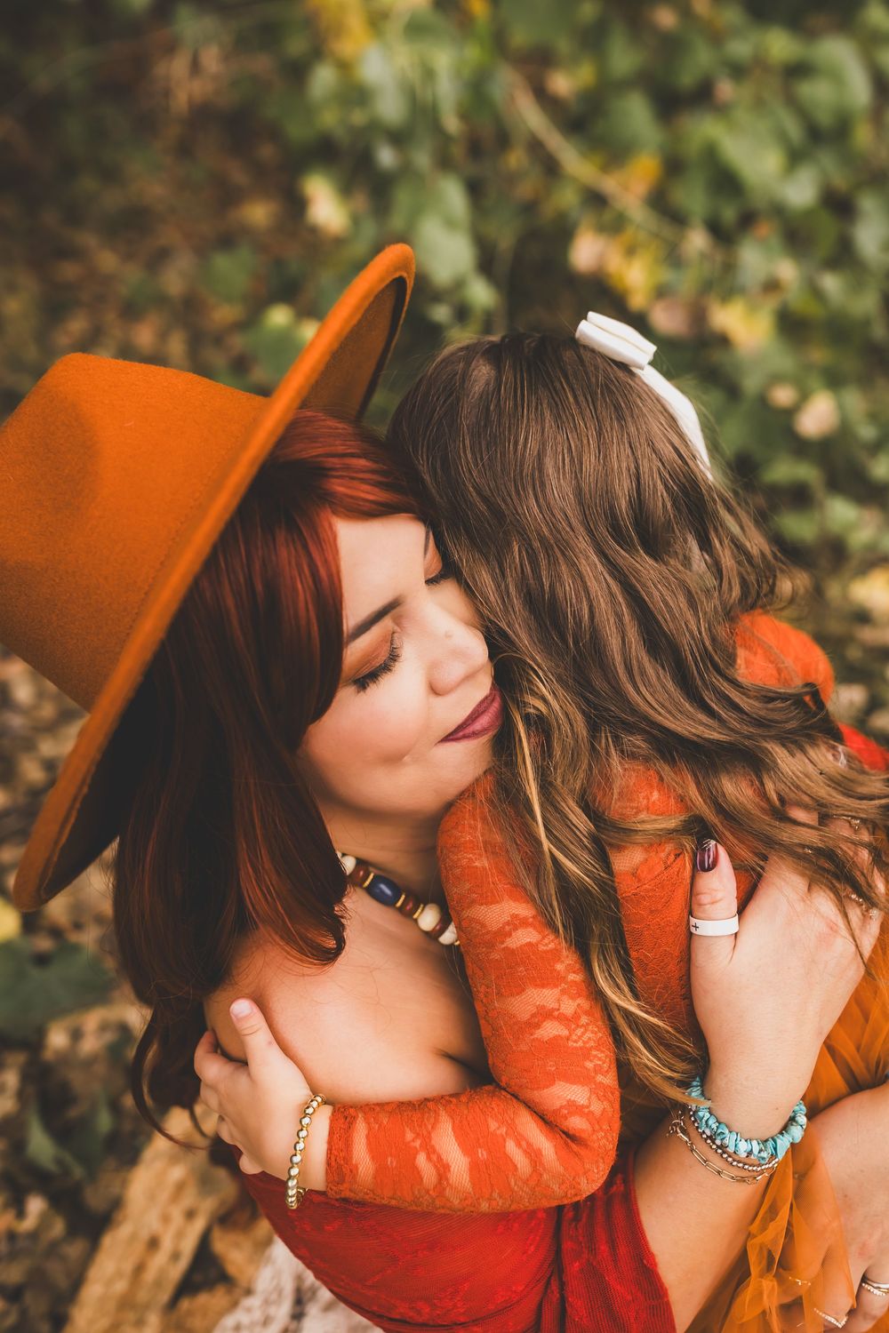 Woman in orange hat and red lace dress tenderly embraces a young girl in an outdoor autumn setting.