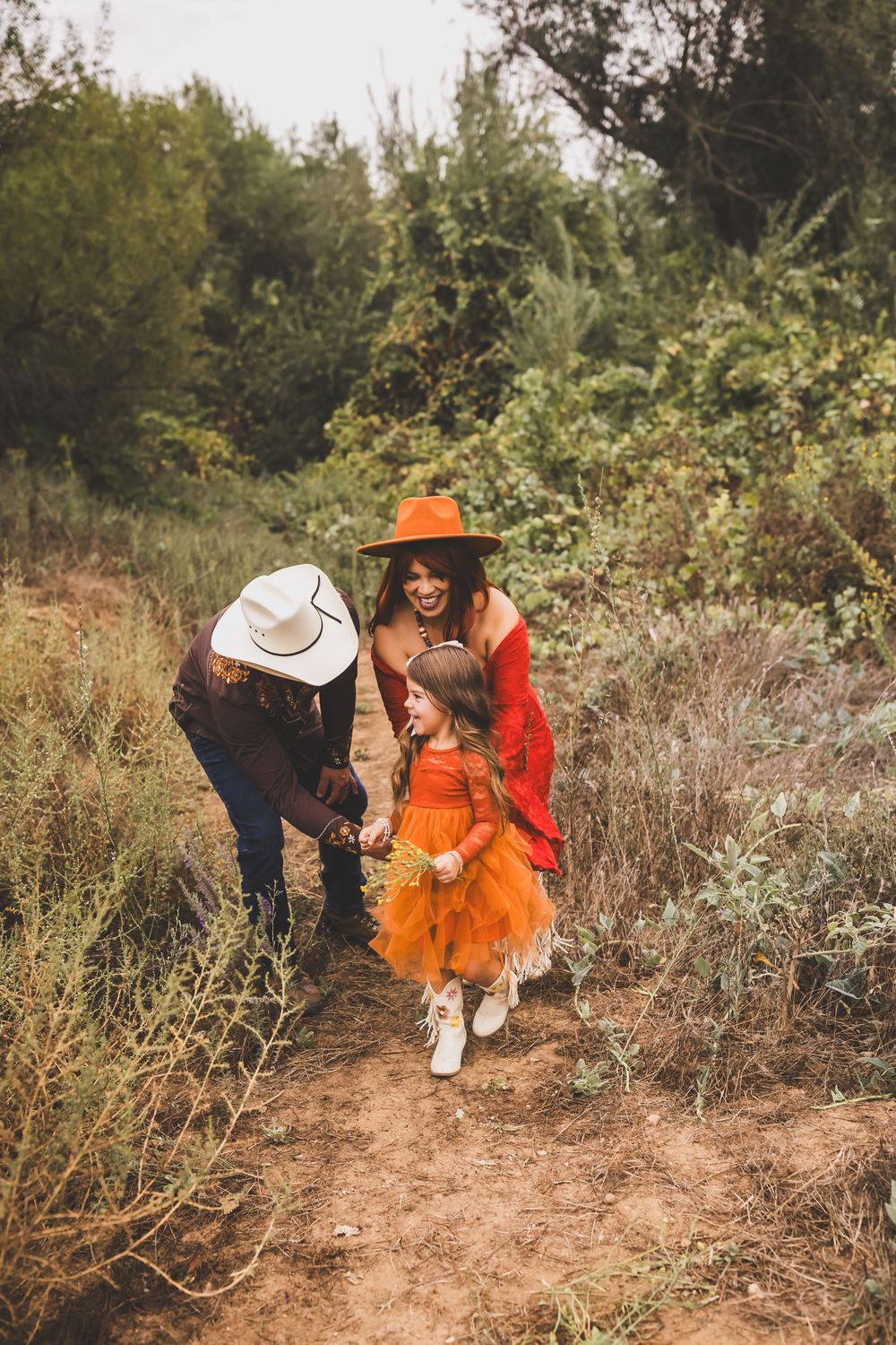 Two women in hats and a young girl in an orange tutu dress walk together on a nature trail surrounded by greenery.