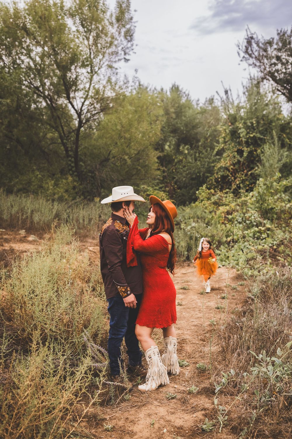 Couple in cowboy hats sharing a kiss in a rustic outdoor setting, with a young girl in an orange dress running toward them.