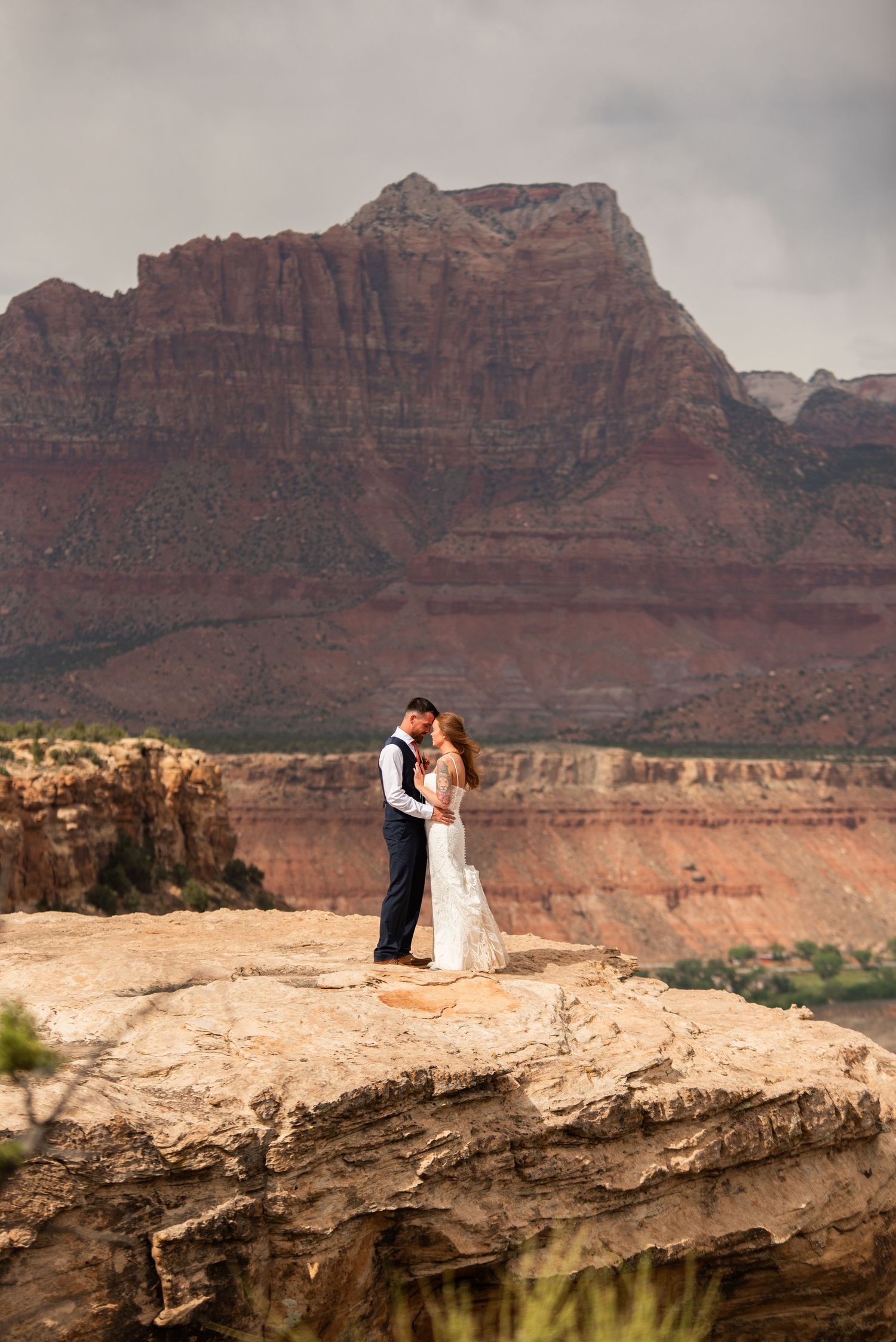 Couple embraces on a rocky cliff edge with stunning red rock canyon formations in Zion National Park backdrop.