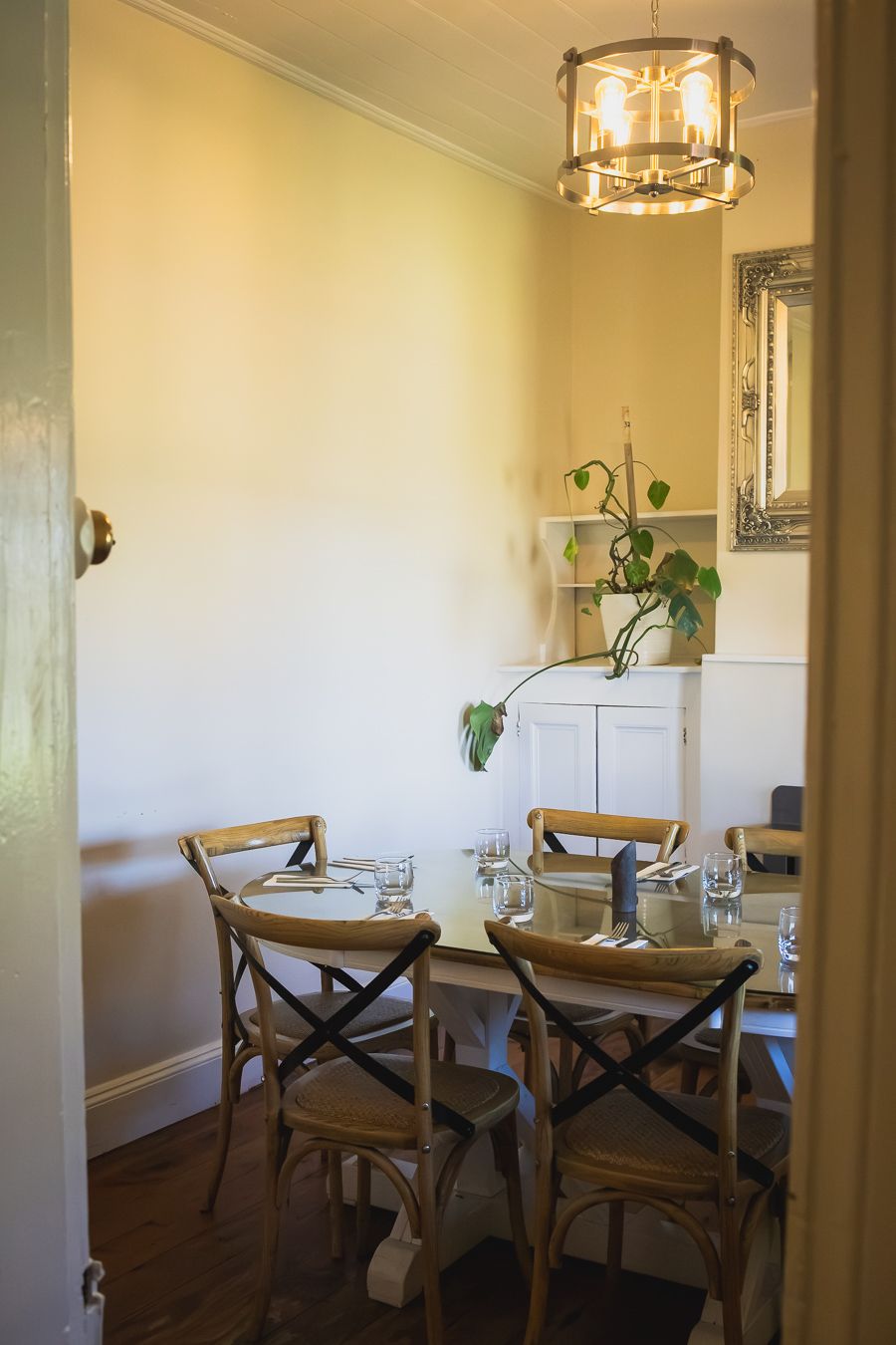 Cozy restaurant dining room with wooden cross-back chairs, set table, pendant light, and green plant on white cabinet.