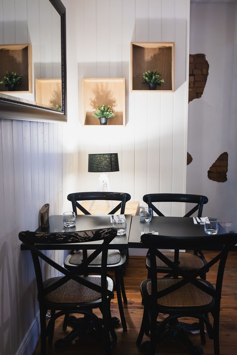 Cozy restaurant corner with black chairs, dark tables, wall-mounted shelves with plants, and a small lamp.
