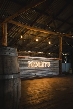Rustic barn venue with glowing Henley's neon sign, string lights, wooden barrel, and corrugated metal walls.