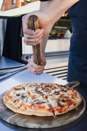 Chef grinding pepper onto a freshly sliced mushroom pizza on a wooden board at a food truck.