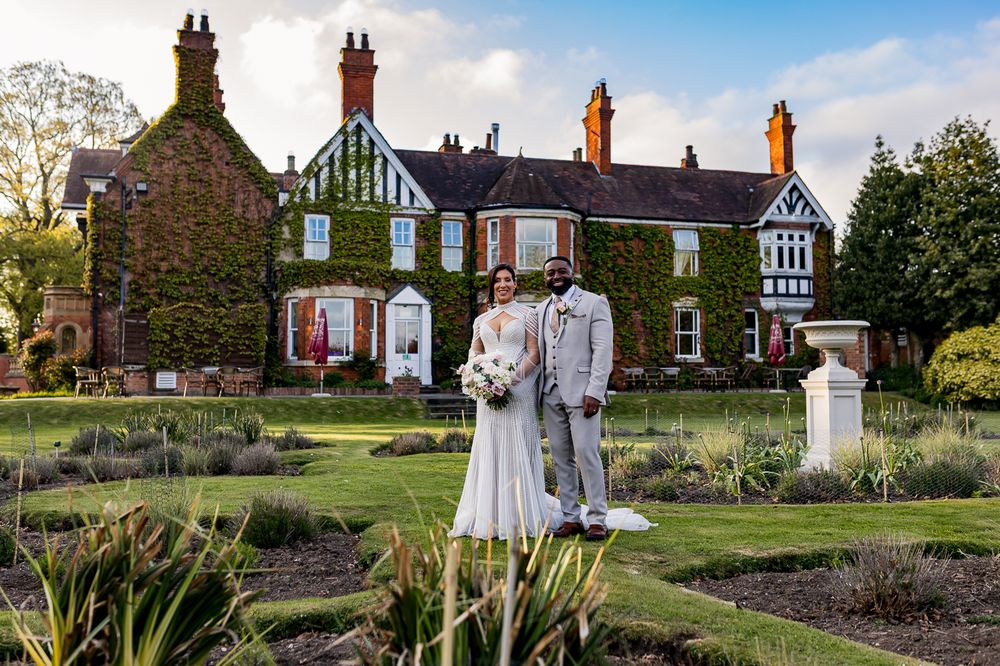 Bride and groom pose in formal garden before ivy-covered Tudor manor house at golden hour wedding.