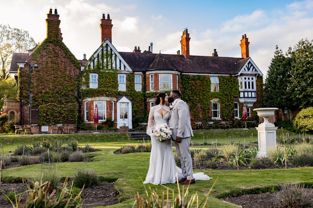 Bride and groom stand in formal gardens before an ivy-covered Tudor manor house at golden hour.