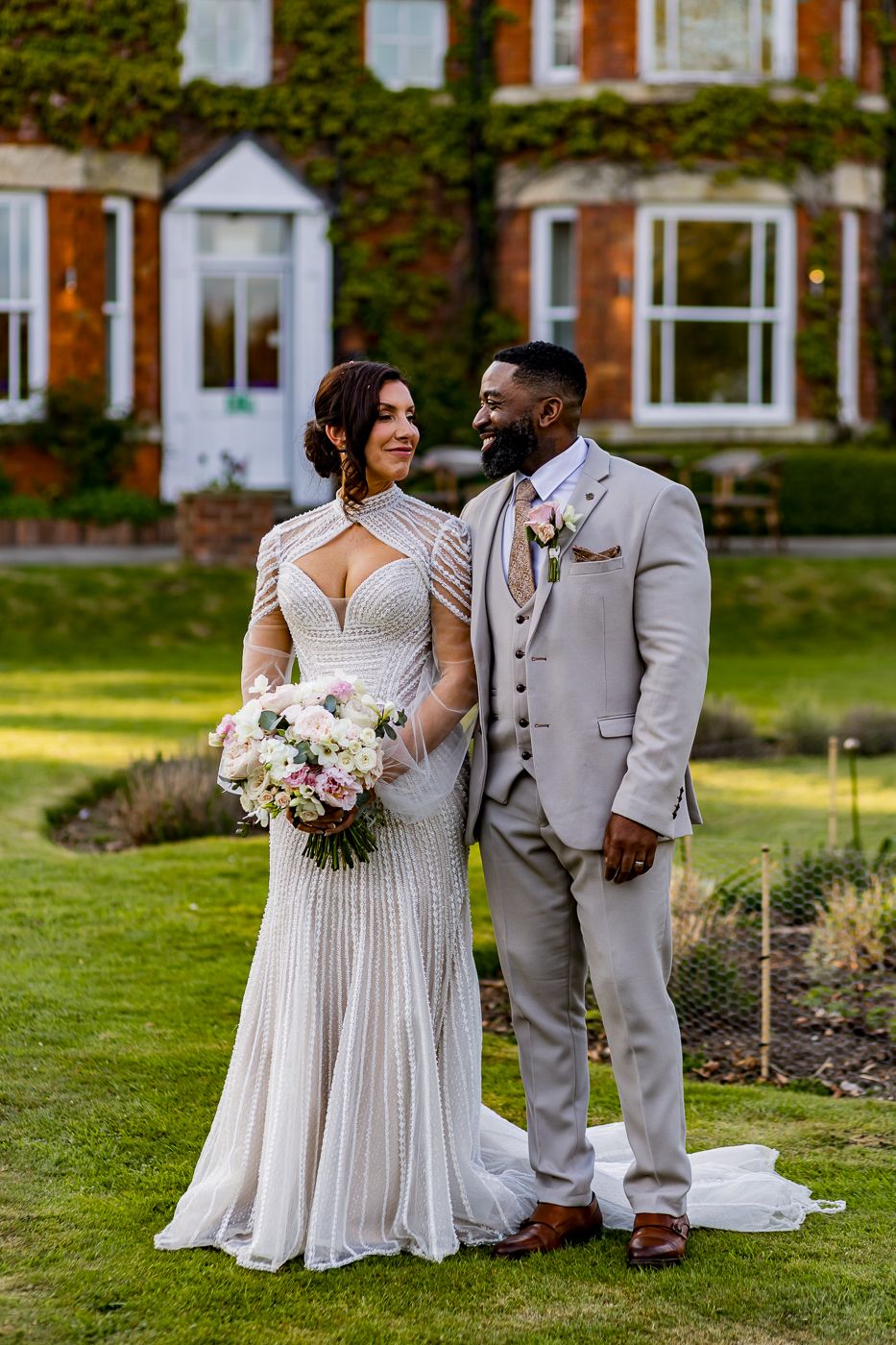 A bride and groom pose elegantly on a green lawn before an ivy-covered brick manor on their wedding day.