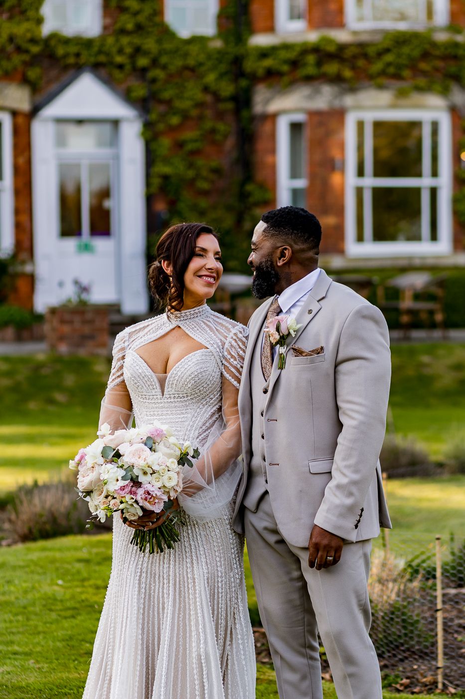 A bride in a beaded gown and groom in a tan suit share a loving glance outdoors at an elegant brick manor.