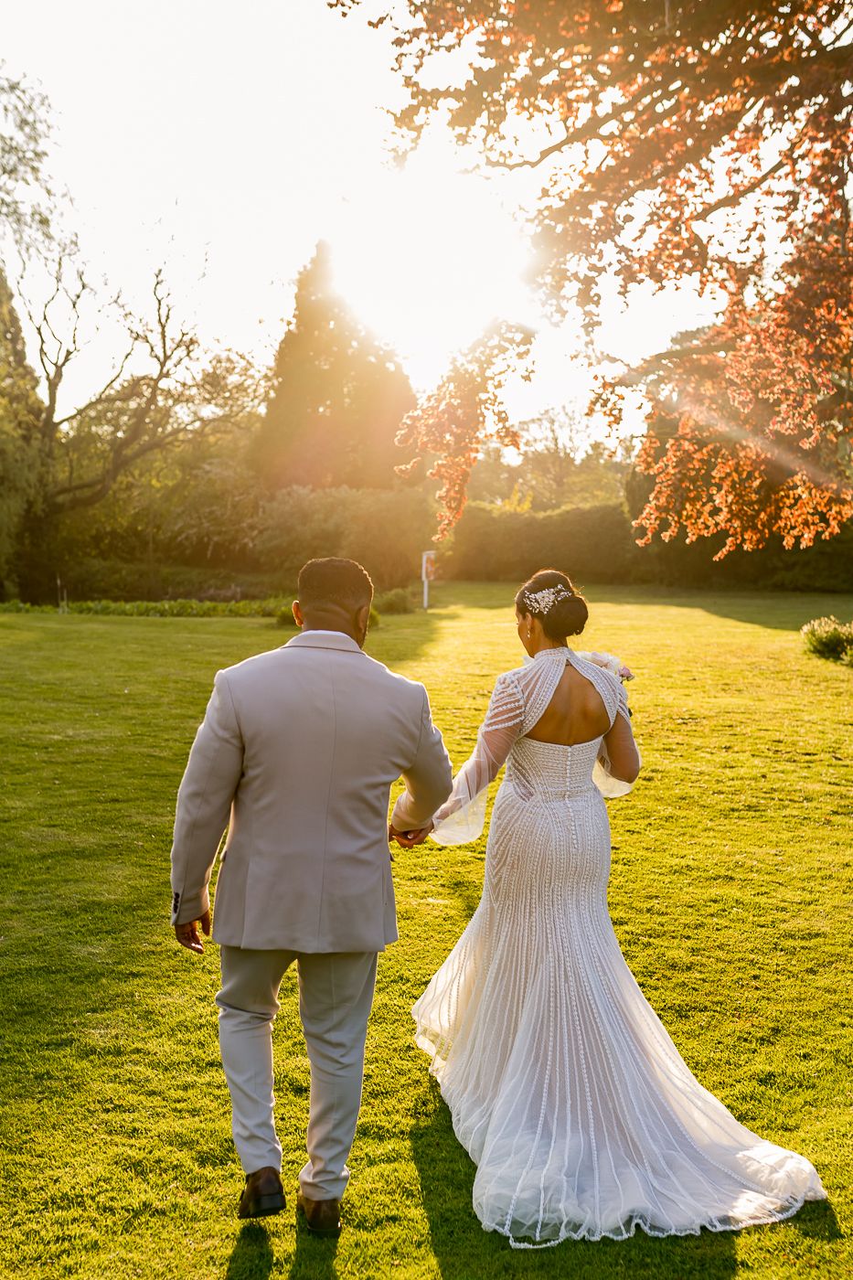 Bride and groom walking hand in hand across lush green lawn at golden sunset, wedding dress with open back and train.