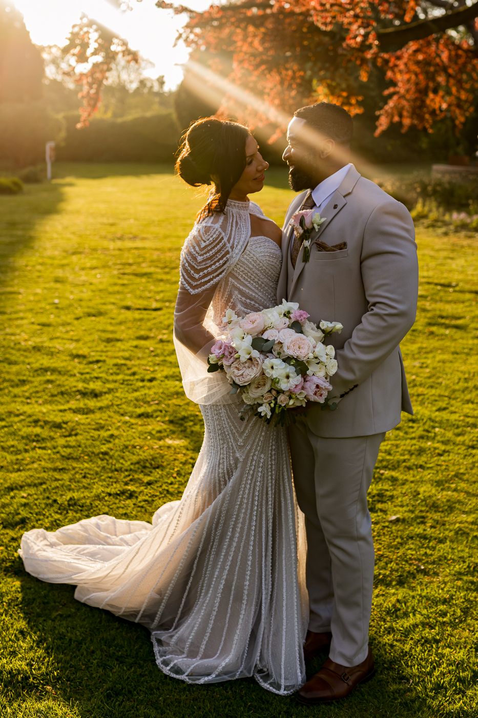 Bride and groom share a romantic moment at golden hour, with sunbeams and autumn foliage in a lush garden setting.