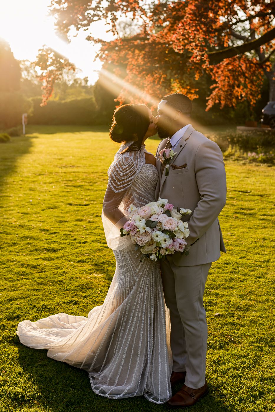Bride and groom kissing at golden hour sunset on lush green lawn, bride in beaded gown holding pink bouquet.