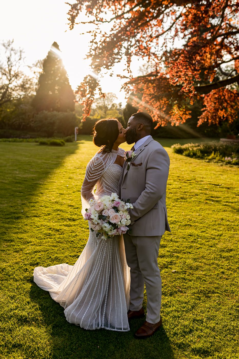 Newlywed couple sharing a kiss at golden hour on lush green lawn, bride in beaded gown holding pink bouquet.