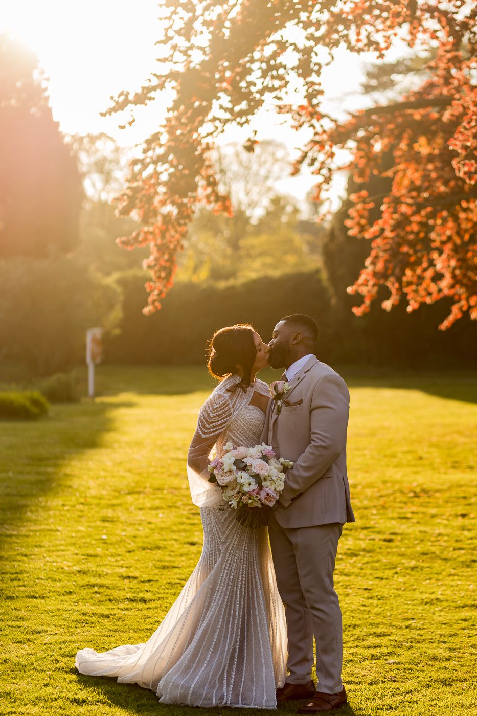 Bride and groom share a kiss at golden hour on lush green lawn with autumn trees glowing in warm sunset light.