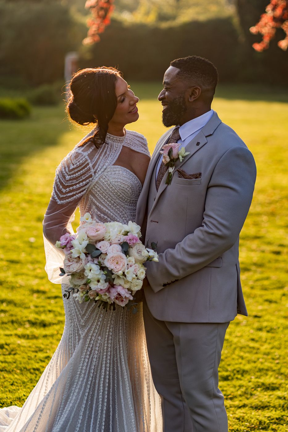 A bride and groom share a romantic moment outdoors at golden hour, she in a beaded gown holding a pink bouquet, he in a tan suit.