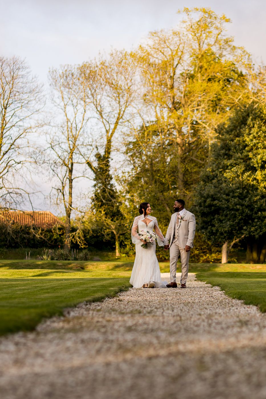 Bride and groom holding hands on a gravel path surrounded by lush green gardens on their wedding day.