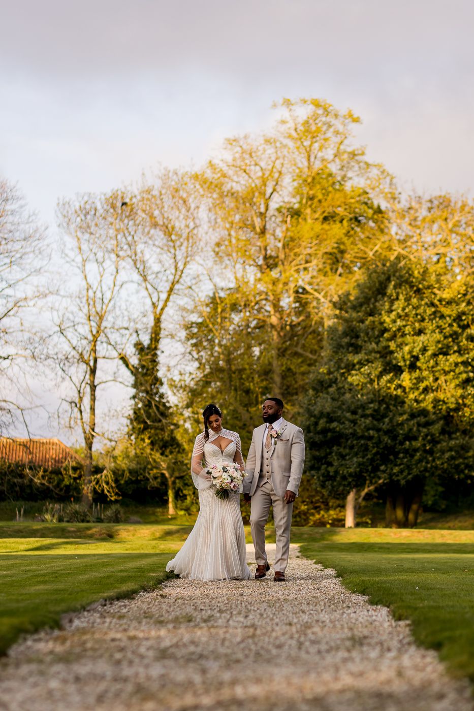 A bride and groom walk hand in hand along a gravel path in a lush garden on their wedding day.