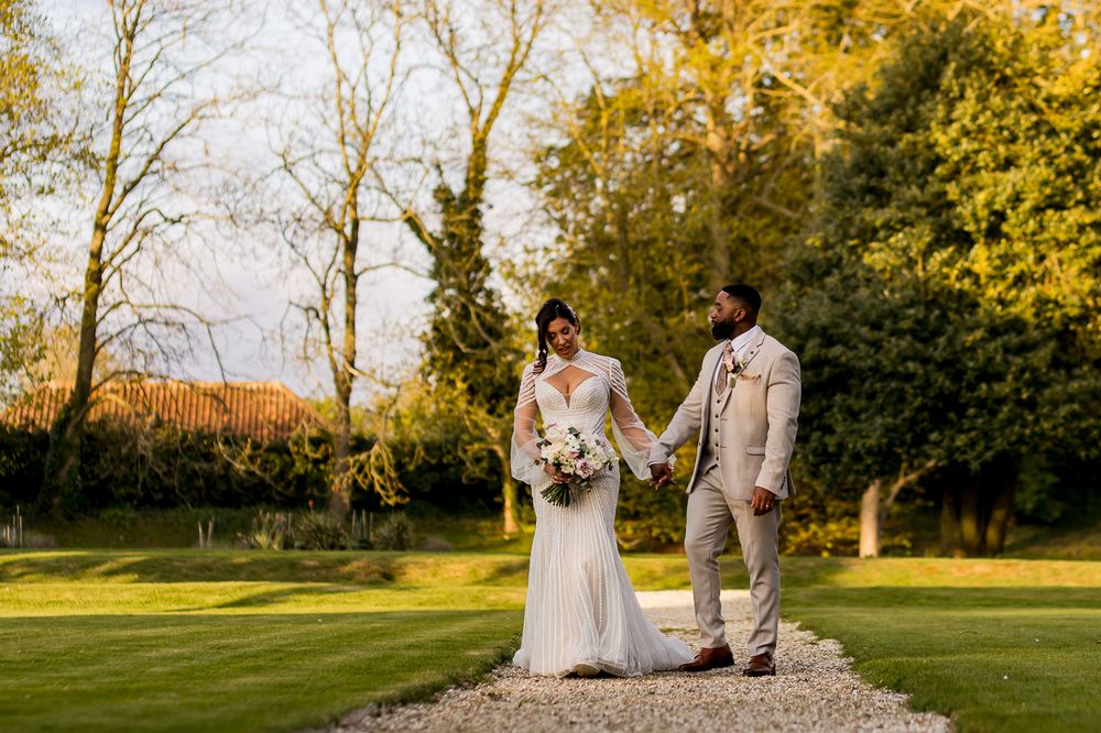 Bride and groom holding hands on a gravel path surrounded by lush green gardens on their wedding day.