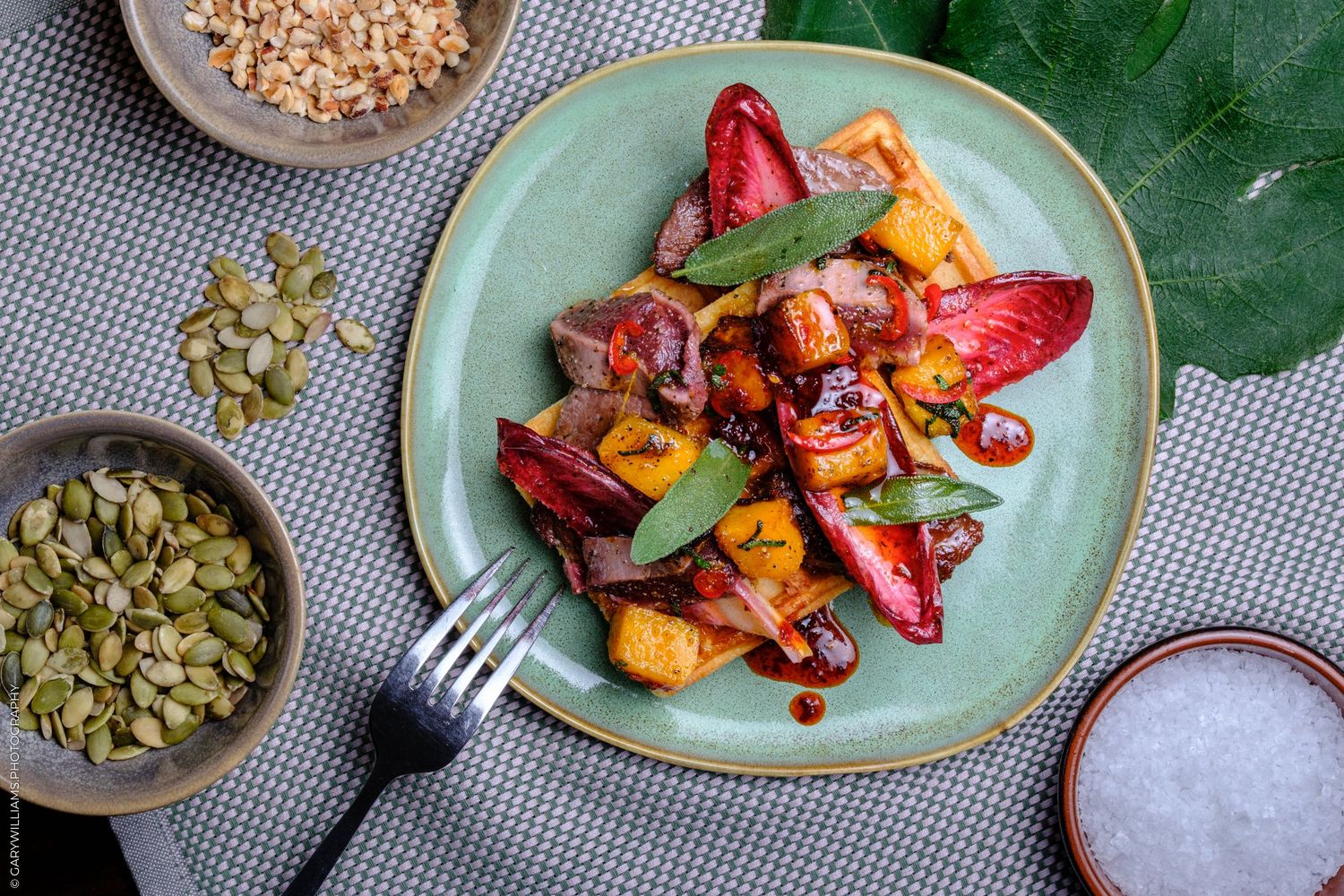 Colorful roasted vegetable salad with strawberries on green plate, surrounded by seeds and salt bowls.