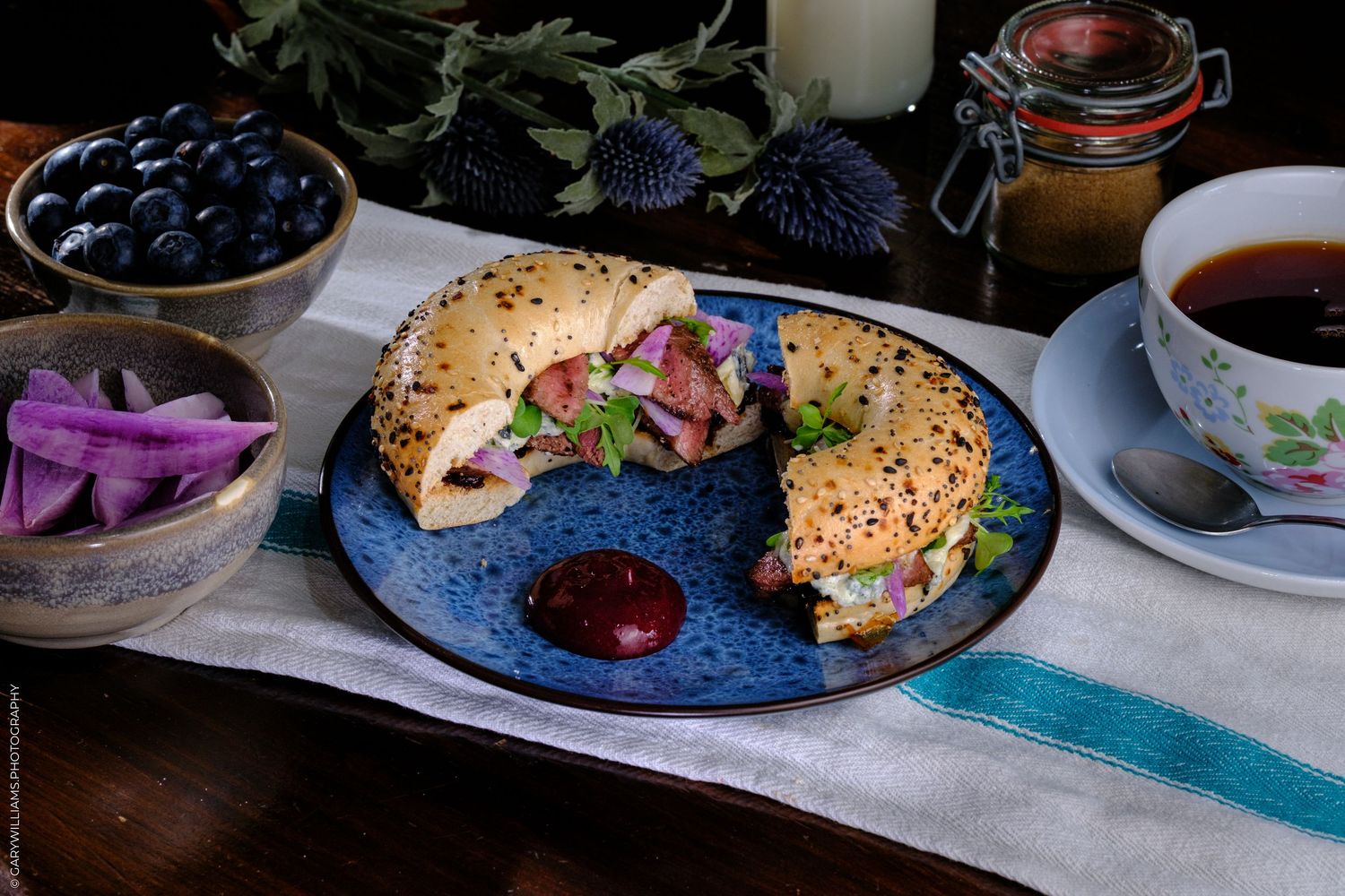 A sliced sandwich on a blue plate with tea, blueberries, and condiments on a rustic table.