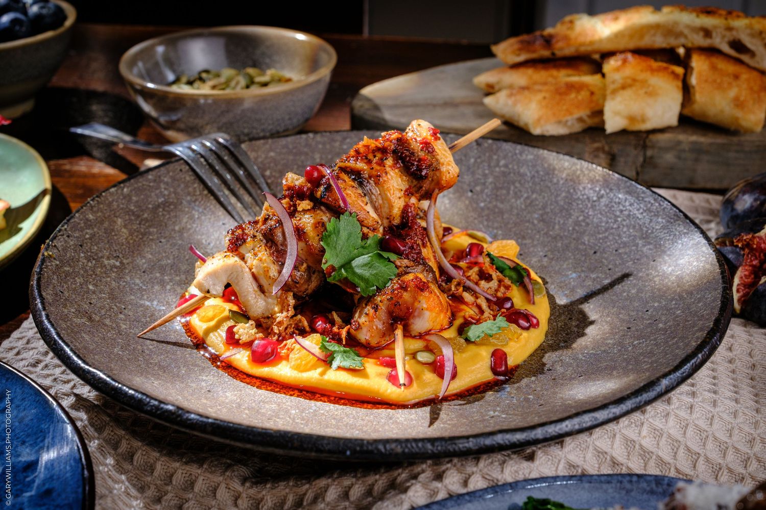 Grilled meat skewers served on creamy hummus with pomegranate seeds and herbs on a rustic dark ceramic plate.