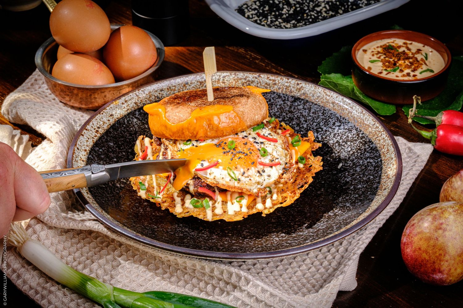 A hand cuts into a loaded breakfast dish with a fried egg, served on a dark plate with fresh vegetables and eggs nearby.