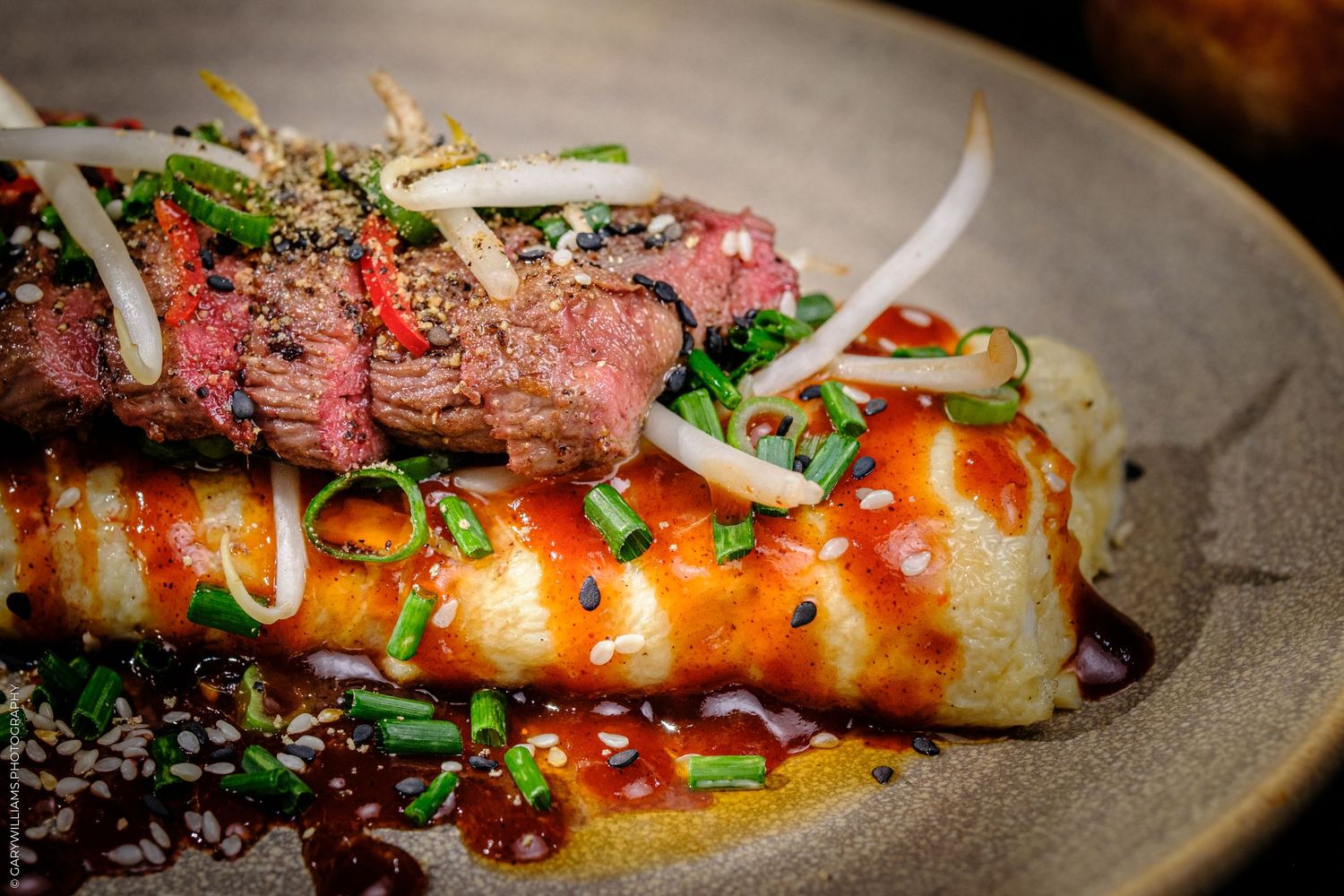 Sliced medium-rare steak served over glazed rice cake, garnished with chives, bean sprouts, and savory sauce.