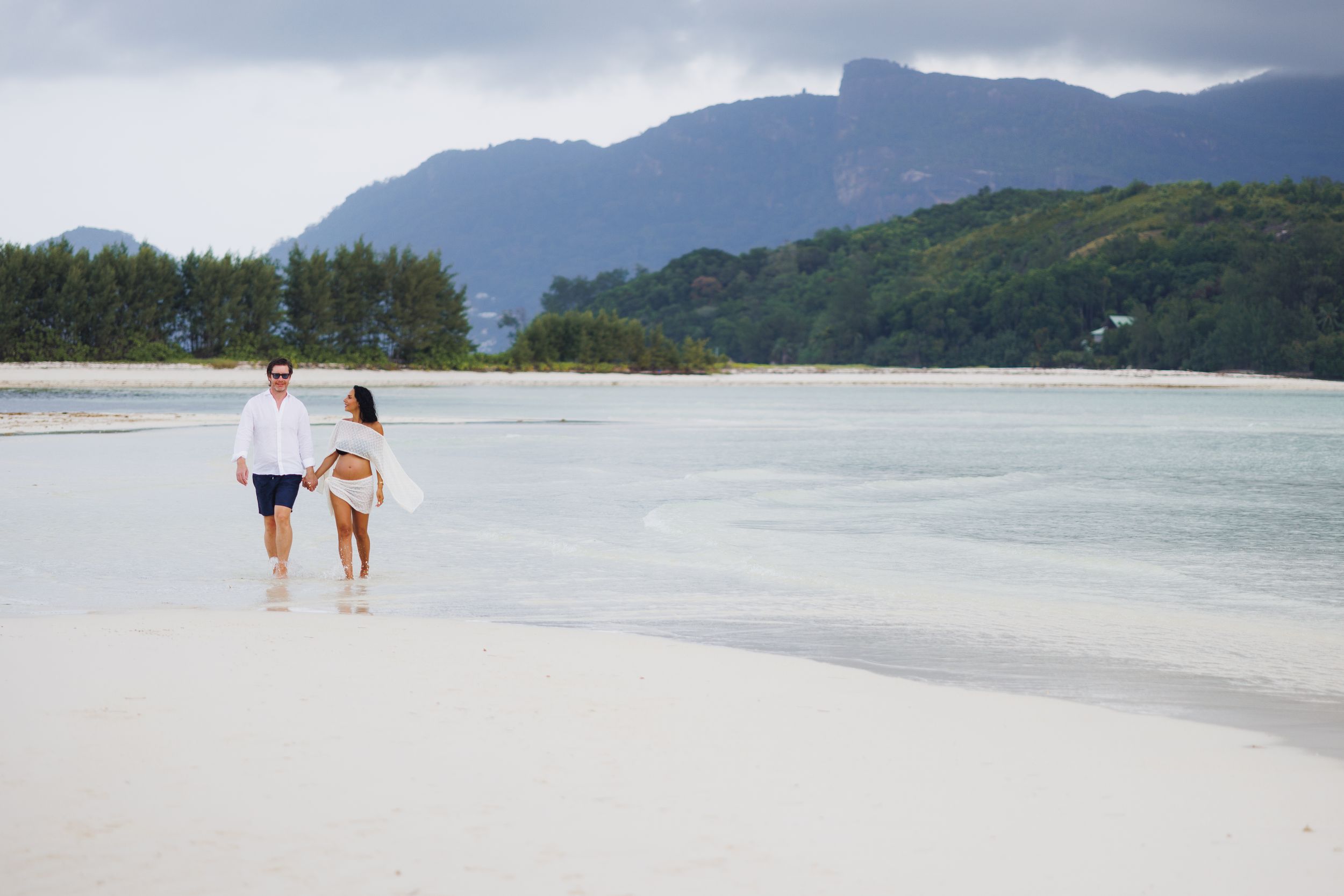 Couple walking hand in hand on pristine white sand beach with turquoise water and mountains.