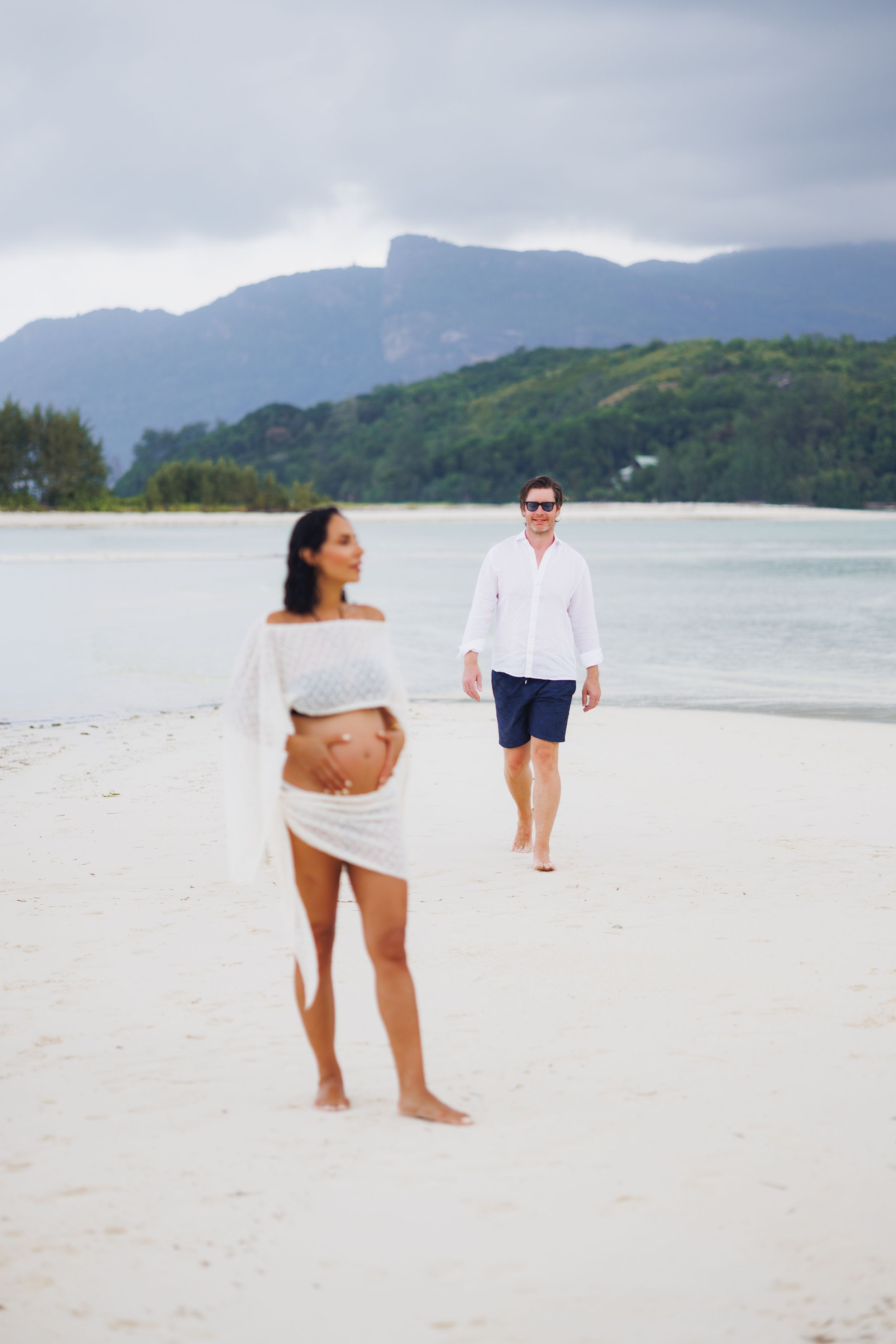 Pregnant woman in white lace top stands on beach as her partner walks toward her with mountains behind.