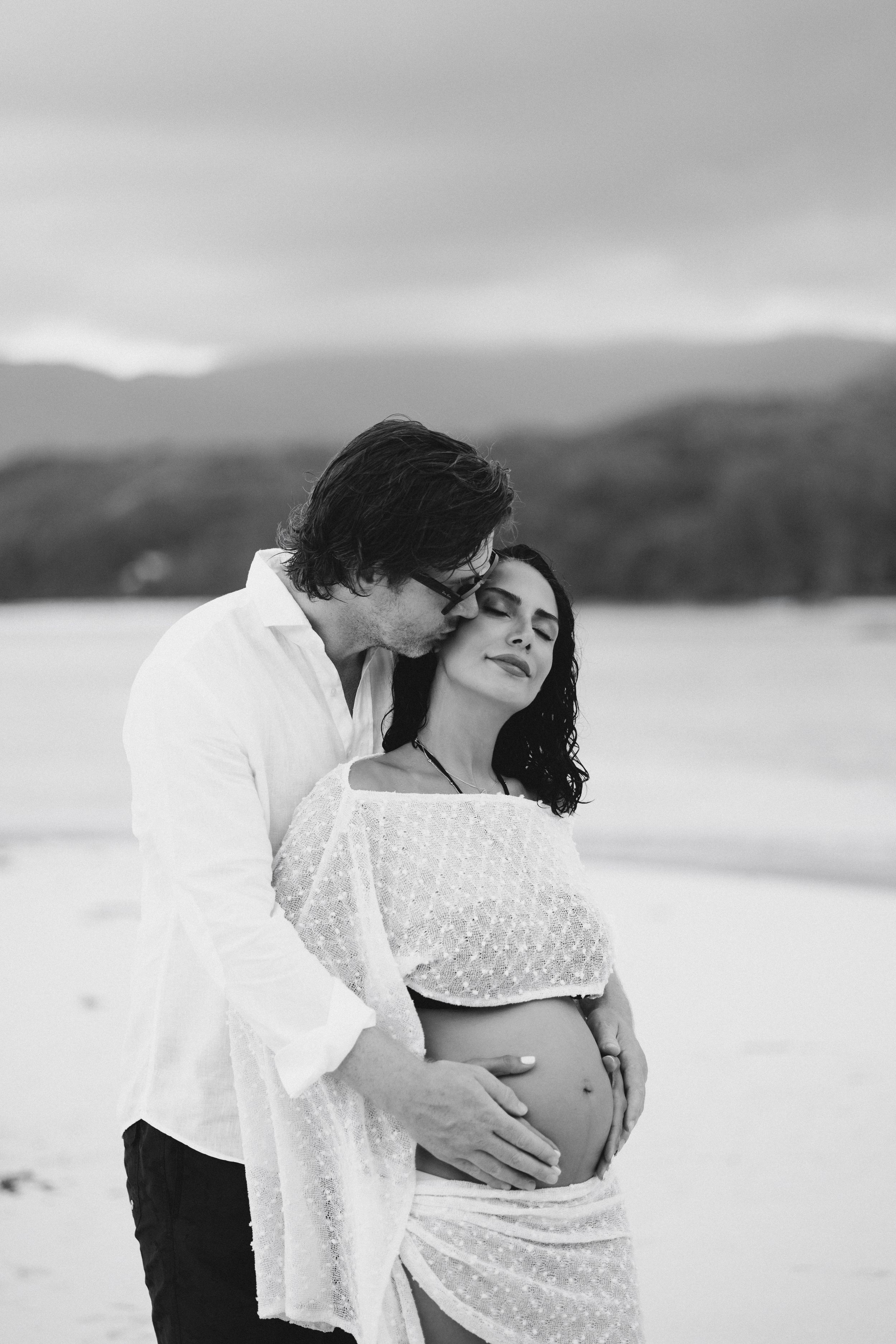 Black and white photo of man kissing pregnant womans head while embracing her belly on beach.