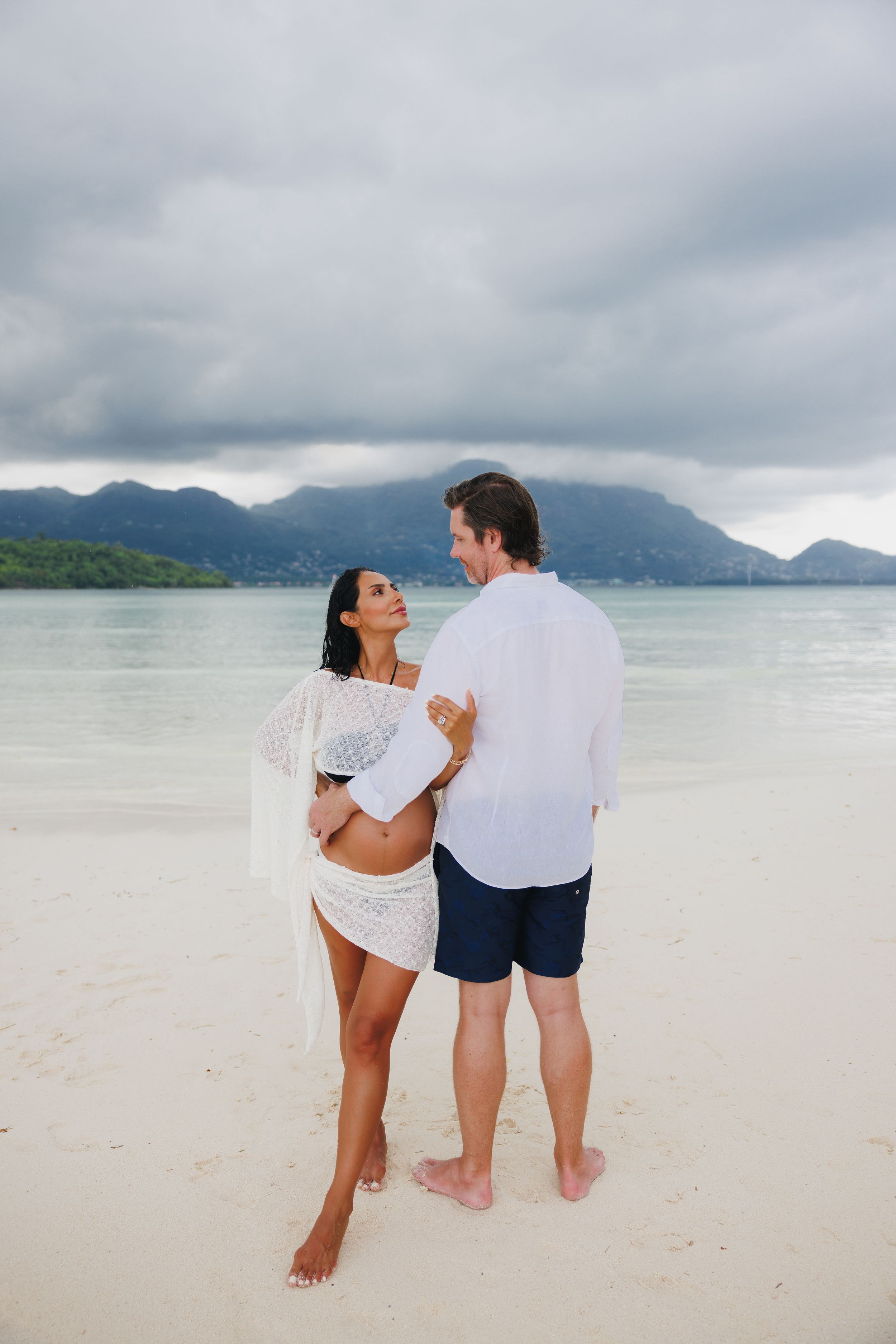 Pregnant couple embracing on a white sand beach with calm turquoise water and mountains in the background.