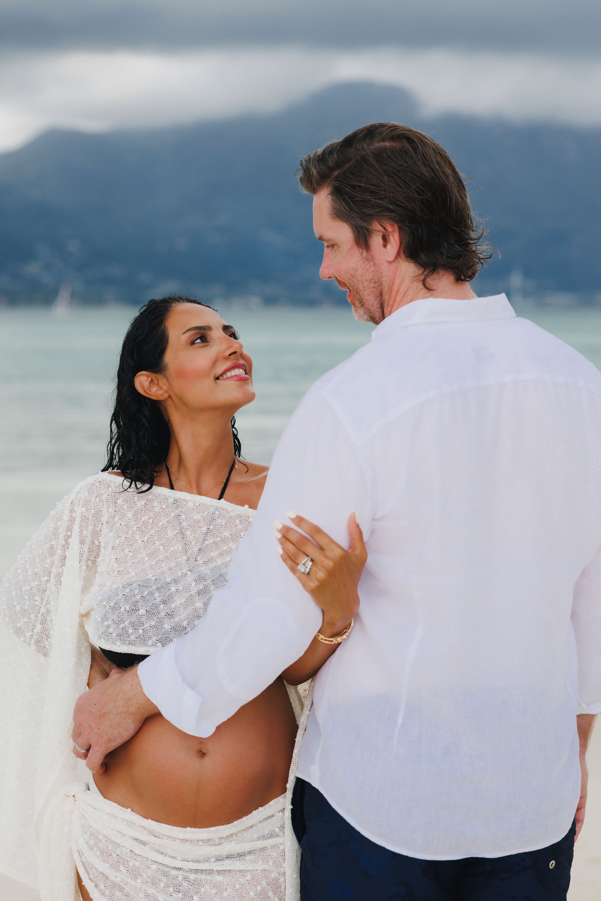 Pregnant woman in white lace wrap smiling at partner on tropical beach with mountains behind.