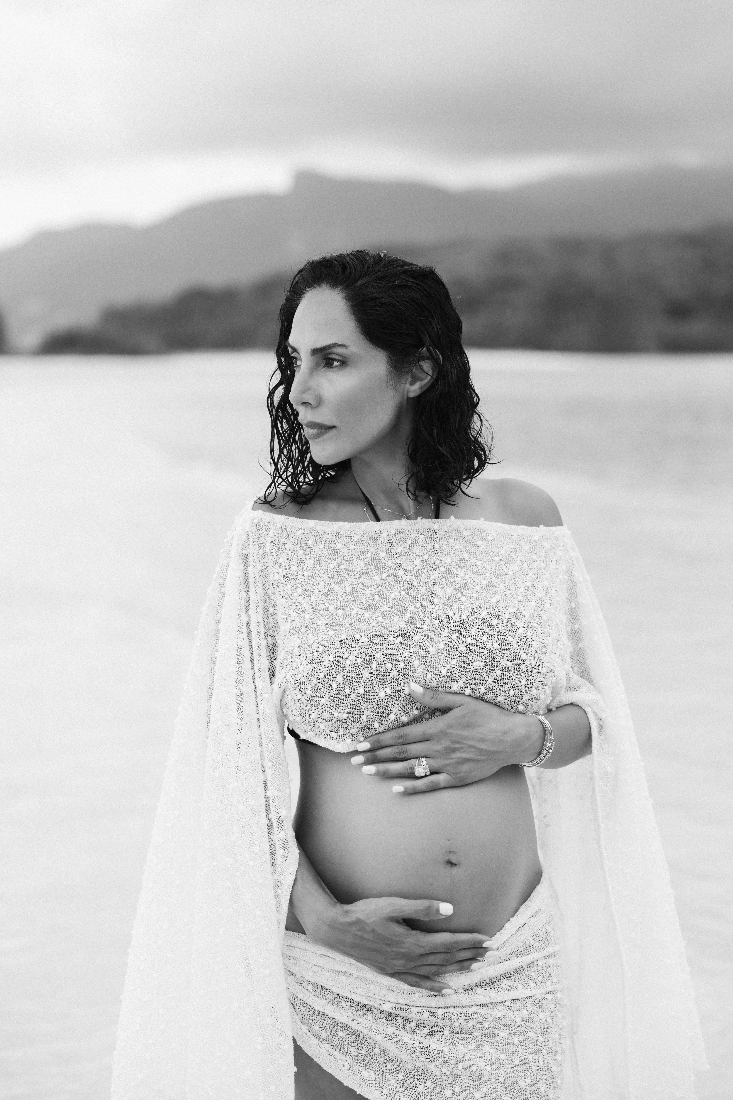 Black and white portrait of a pregnant woman in a white lace dress cradling her baby bump on a beach.