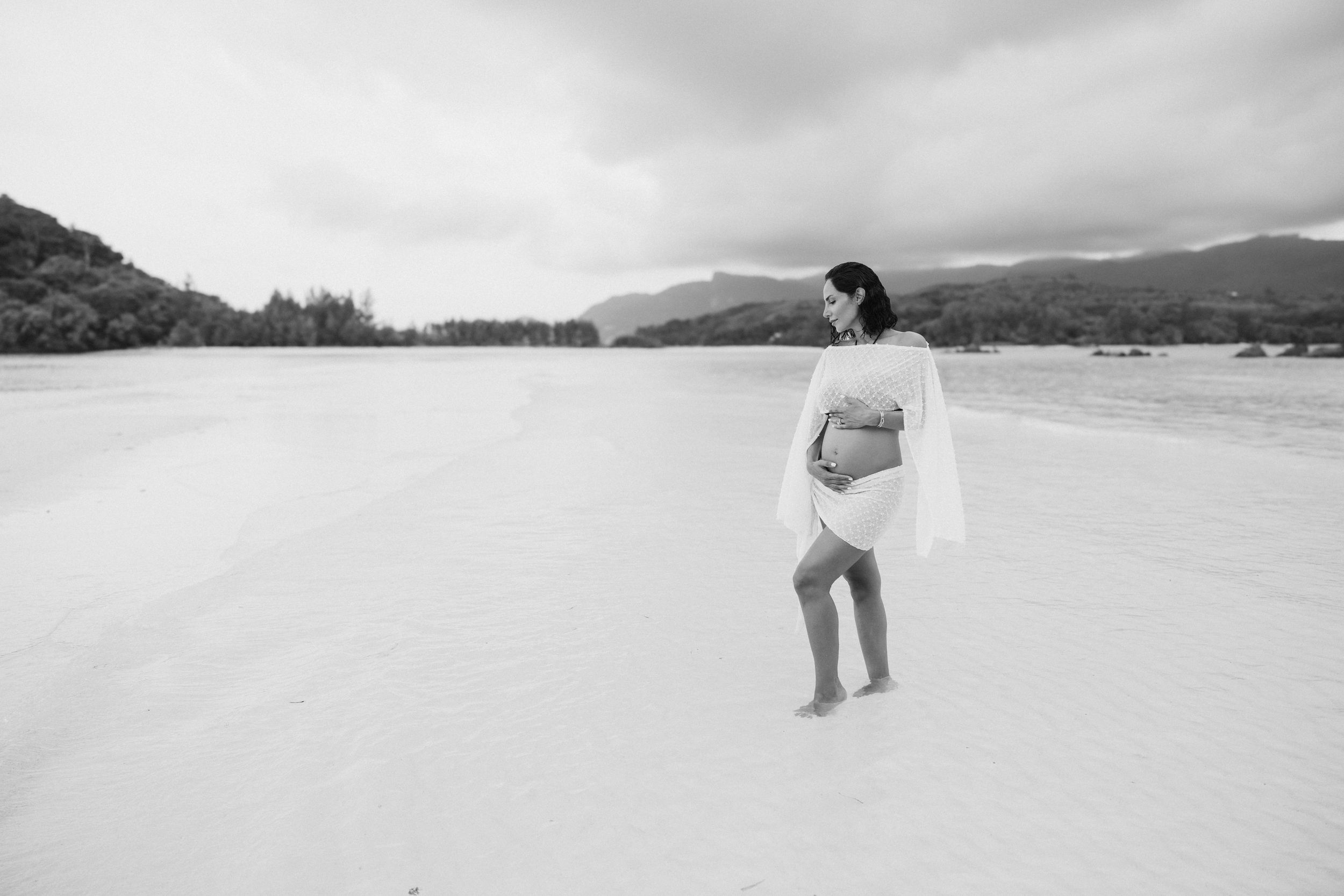 Black and white image of a pregnant woman in a white lace outfit standing alone on a vast sandy beach.