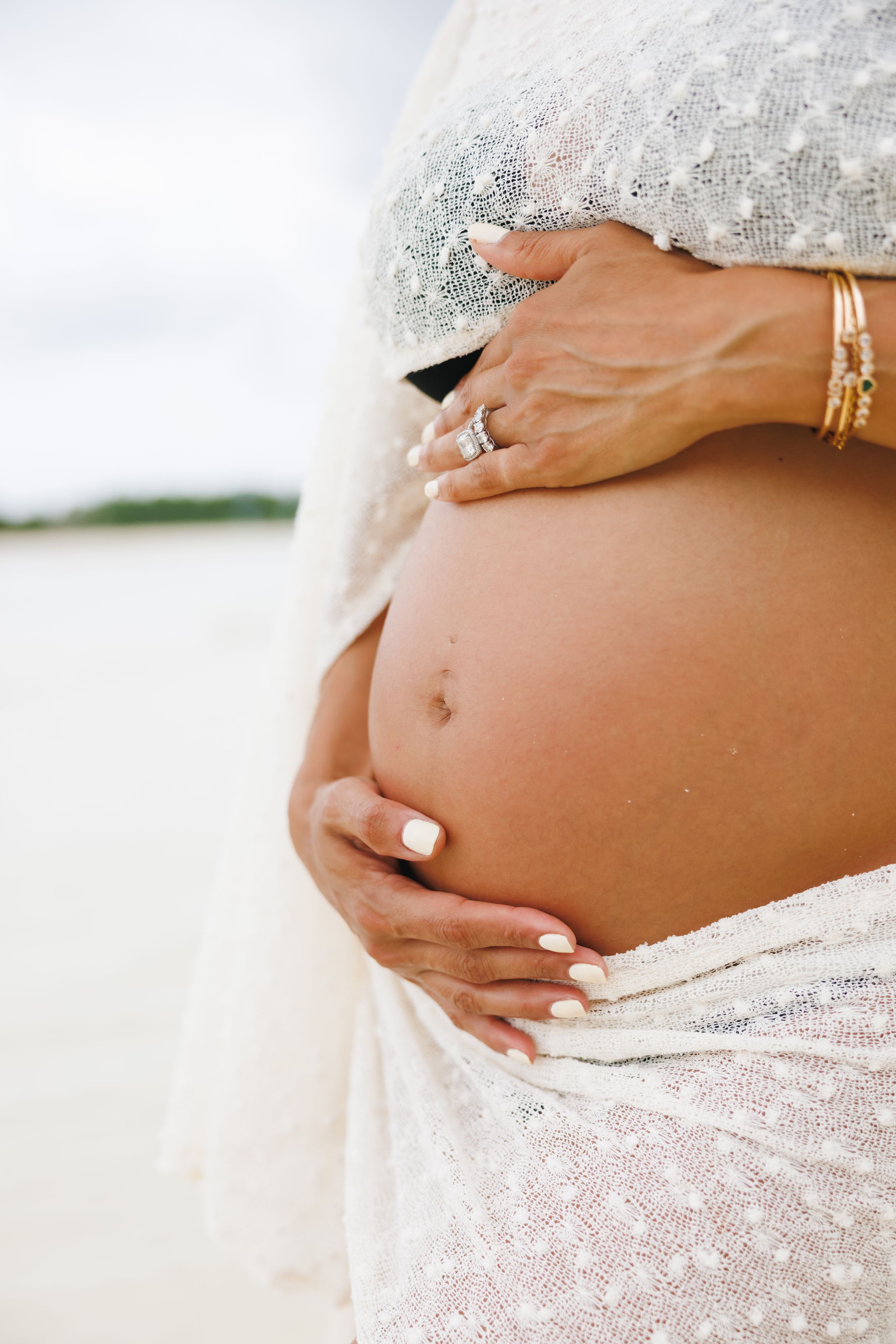 Close-up of a pregnant woman's baby bump with hands adorned with gold bracelets and rings on a beach.