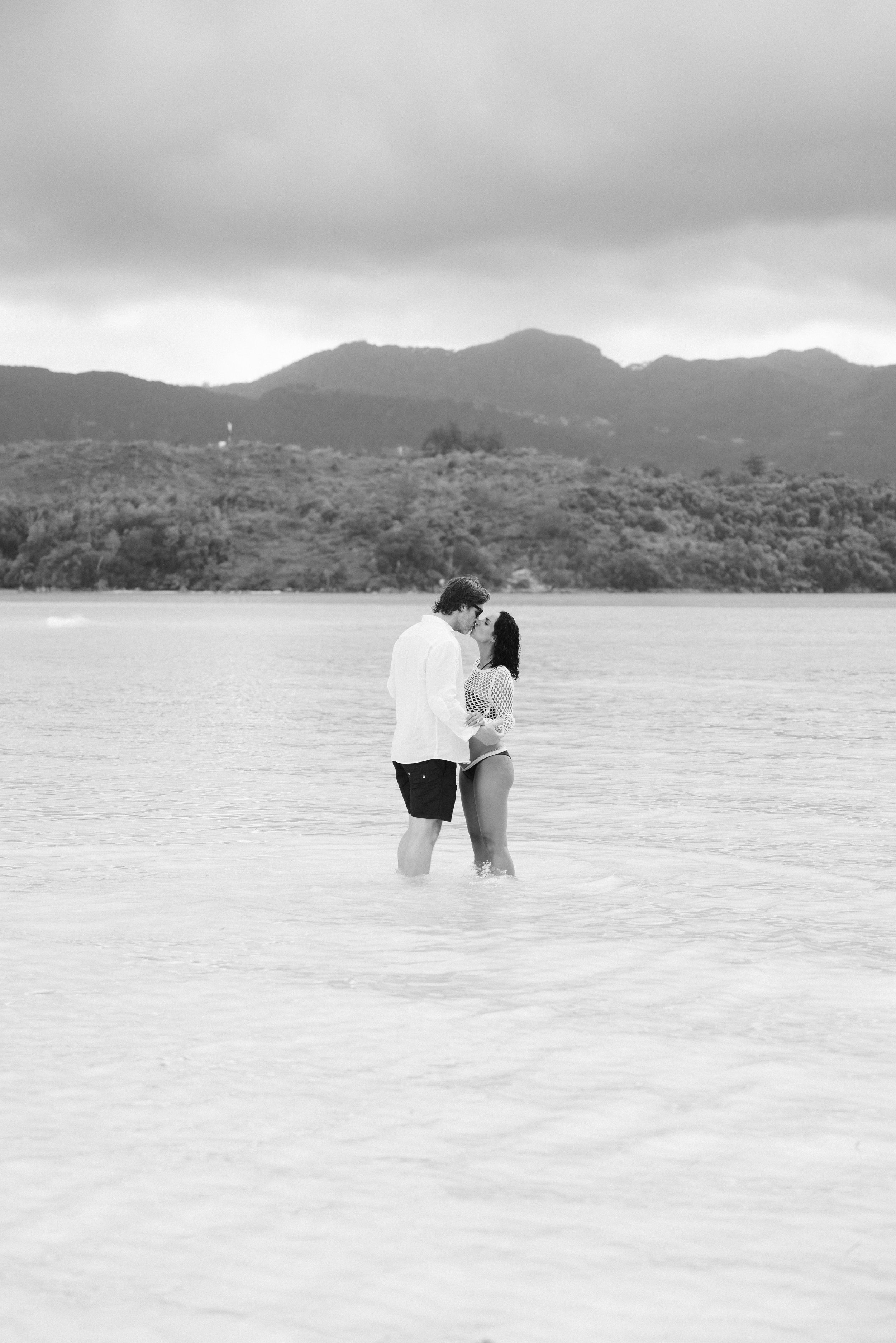 Black and white photo of a couple sharing a romantic kiss while standing in shallow ocean water.