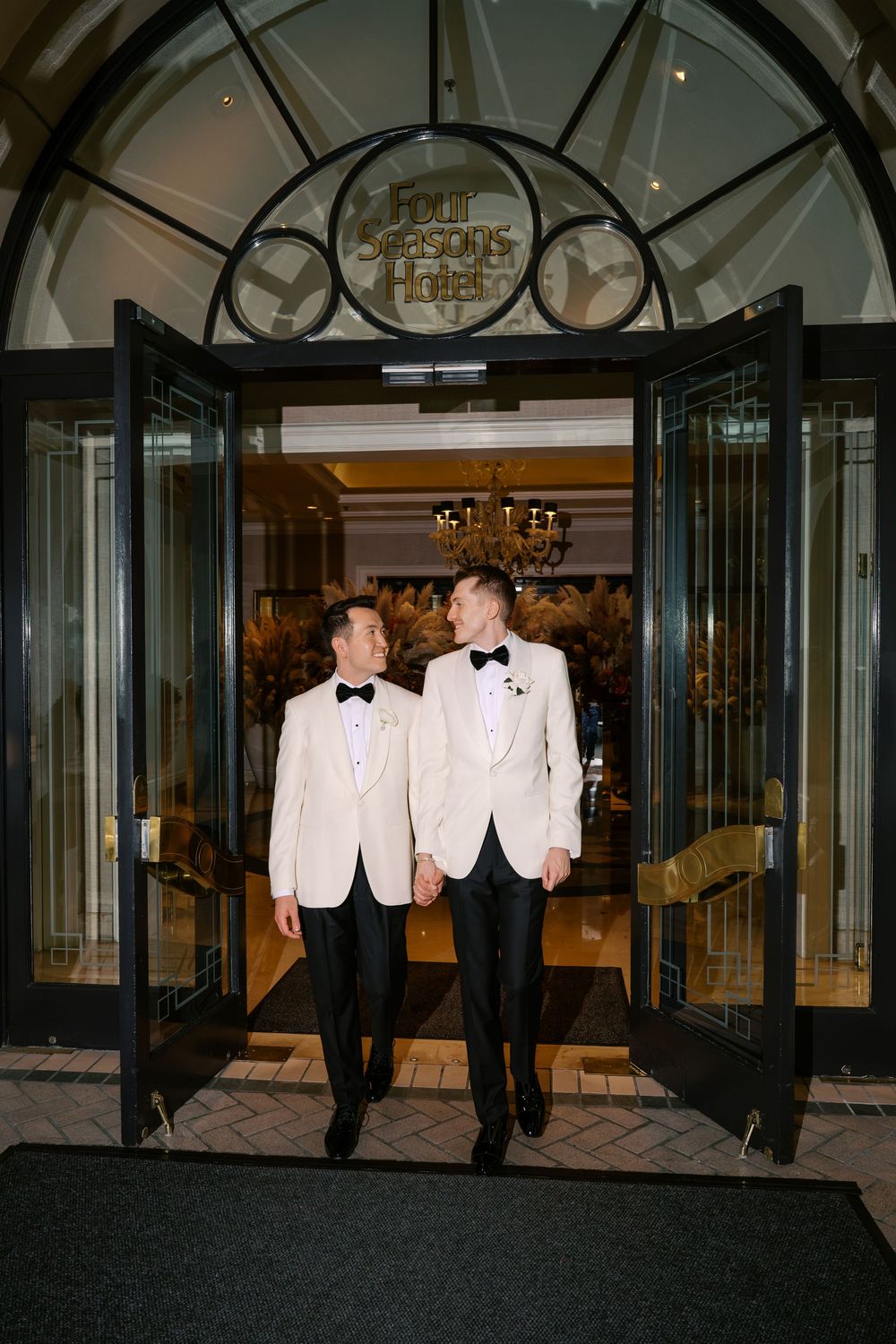 Two grooms in white tuxedo jackets exit Four Seasons Hotel hand in hand on their wedding day.