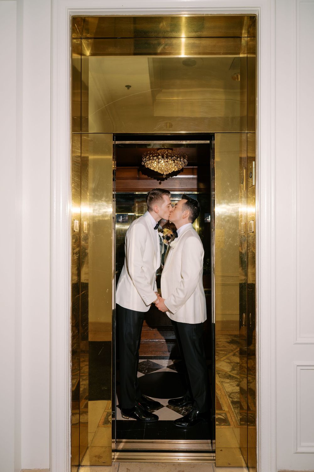 Two grooms in white tuxedo jackets share a kiss inside a gold mirrored elevator at their wedding venue.