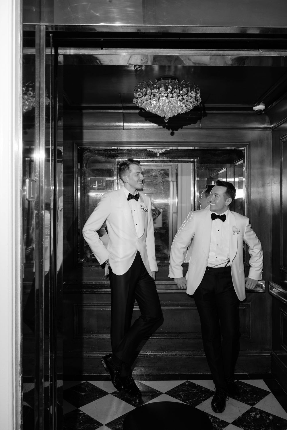 Two grooms in white tuxedo jackets and black bow ties laugh together in an elegant mirrored elevator.