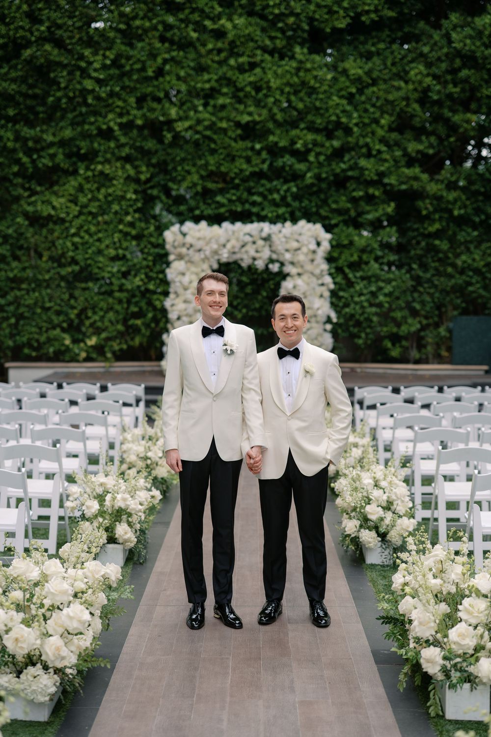 Two grooms in white tuxedo jackets hold hands at an elegant outdoor wedding ceremony with white floral arrangements.