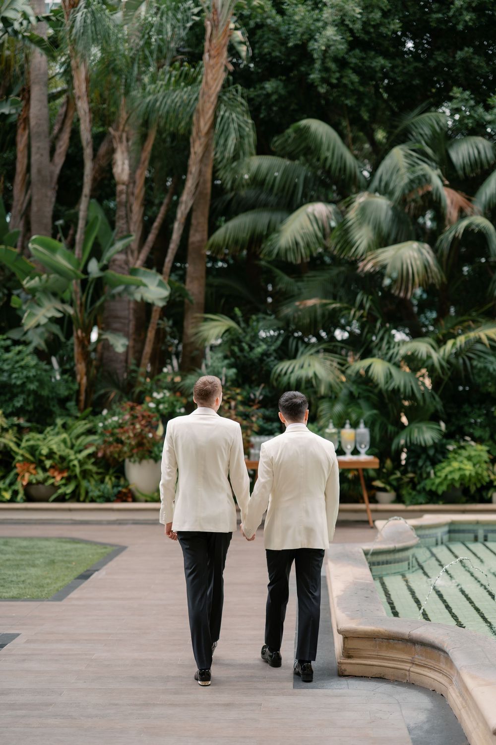 Two grooms in white tuxedo jackets hold hands walking through a lush tropical garden on their wedding day.