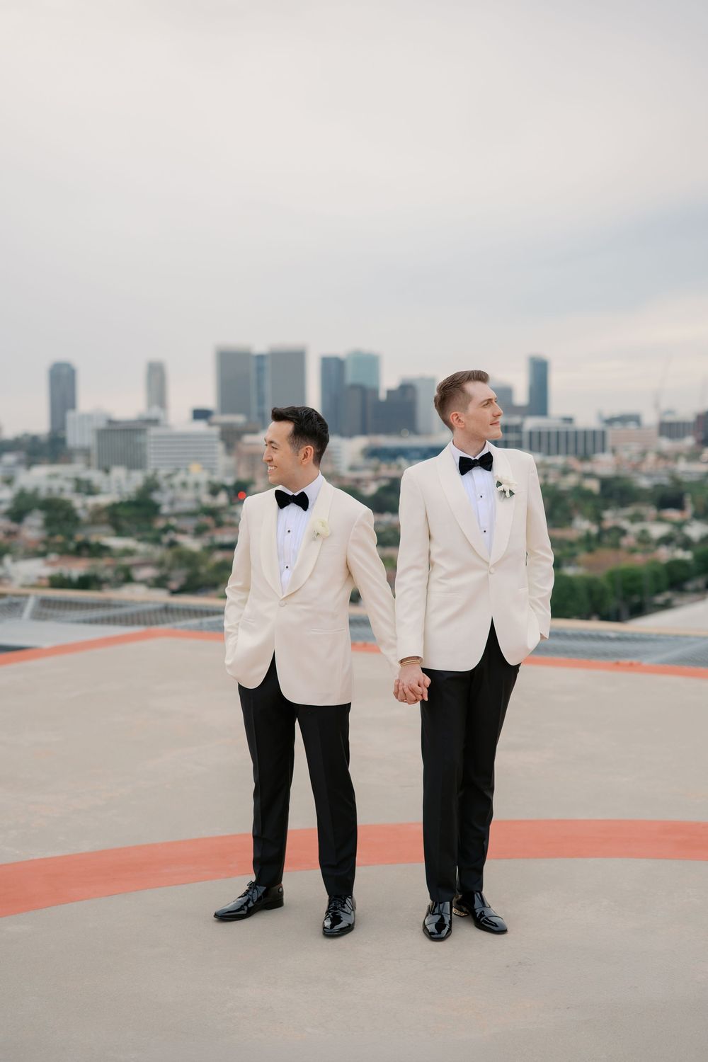 Two grooms in white tuxedos hold hands on a rooftop helipad with a city skyline backdrop.