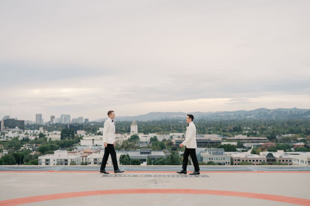 Two grooms in white jackets walk on a rooftop helipad with a city skyline backdrop on their wedding day.