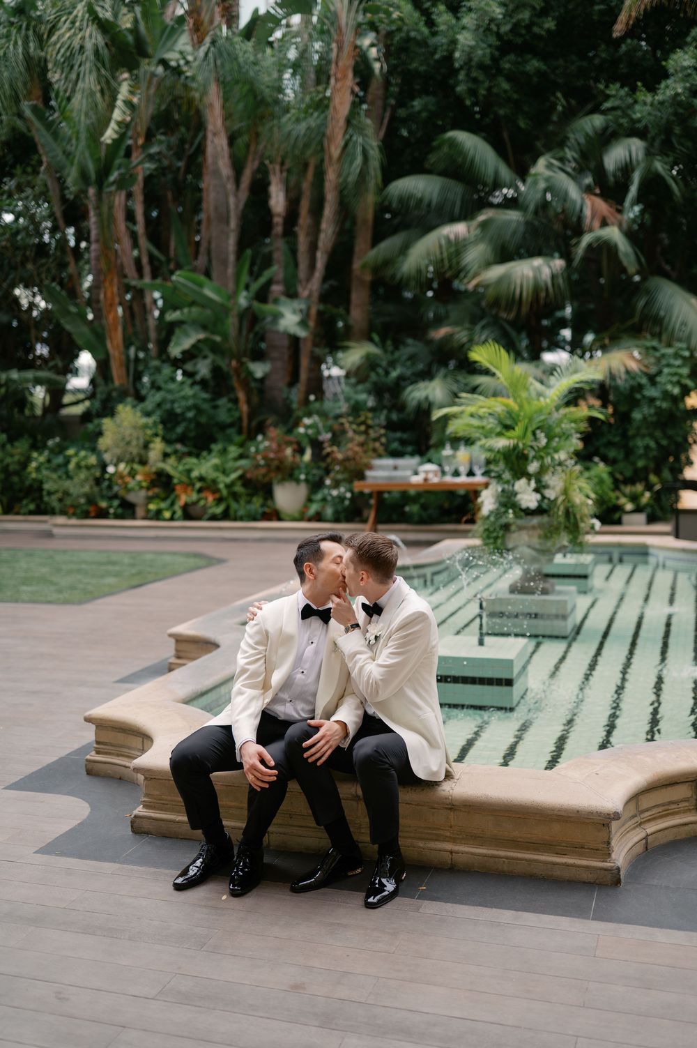 Two grooms in white tuxedo jackets share a kiss beside a fountain surrounded by lush tropical greenery.