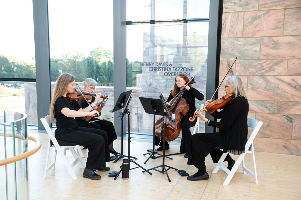 String quartet performing chamber music in a modern venue with large windows, musicians seated with violins and cello.
